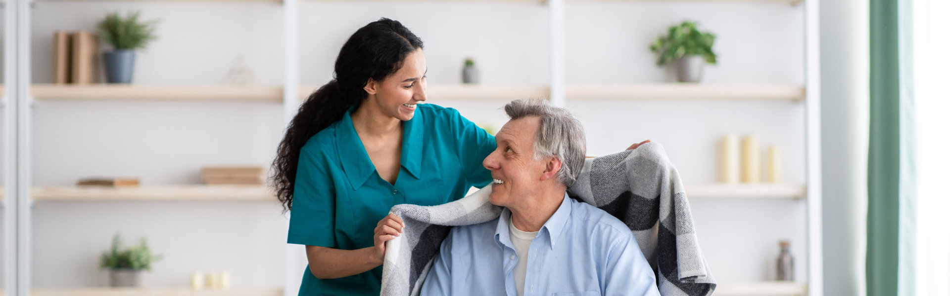 A female caregiver giving a coat to an elderly man sitting on a wheelchair